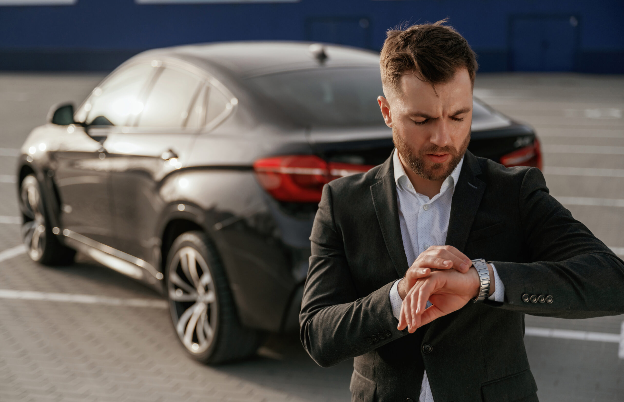 Checks the time. Businessman in suit is near his black car outdoors.